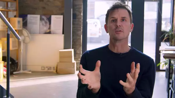 The artist Luke Jerram sits in his studio with boxes, natural light and plants visible behind him.