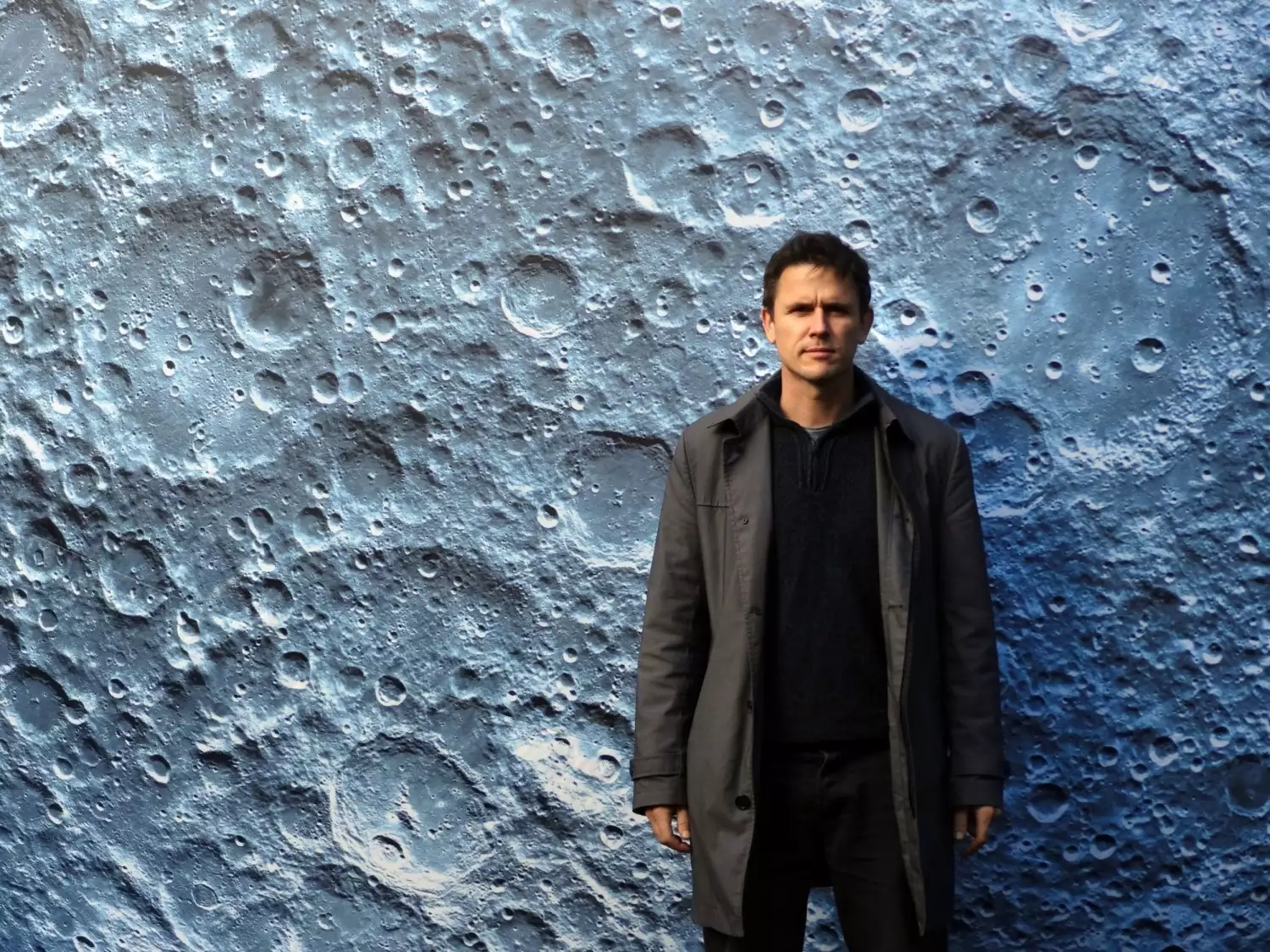 Luke Jerram, a male artist stood in front of a close up of a moon crater artwork