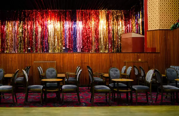 A pub stage with a glittery curtain, blue chairs, tables and a patterned carpeted floor.